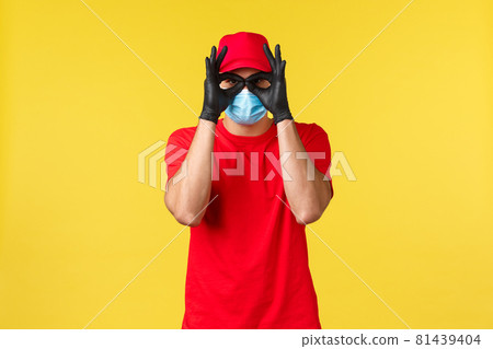 Express delivery during pandemic, covid-19, safe shipping, online shopping concept. Funny courier in red uniform, medical mask and gloves, show okays or glasses with hands 81439404