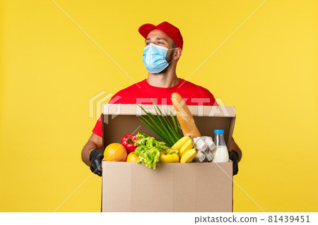 Takeaway delivery, covid-19 quarantine and groceries concept. Carefree handsome courier in red uniform and medical mask, look around with smile, holding box with food order 81439451
