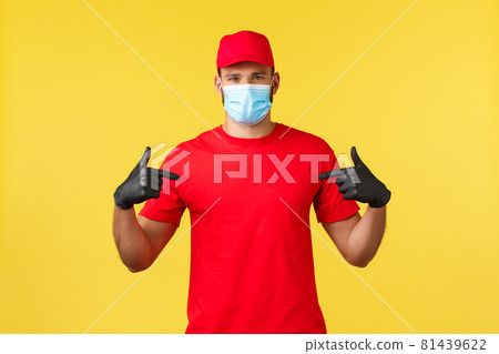 Express delivery during pandemic, covid-19, safe shipping, online shopping concept. Young courier in red uniform, medical mask and gloves pointing at himself, provide fast delivery of packages 81439622