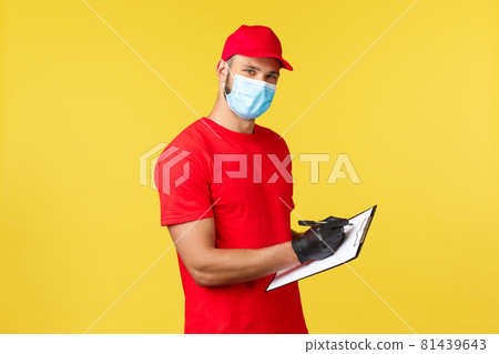 Express delivery during pandemic, covid-19, safe shipping, online shopping concept. Pleasant young courier in red uniform and medical mask, fill in order on clipboard, look camera 81439643