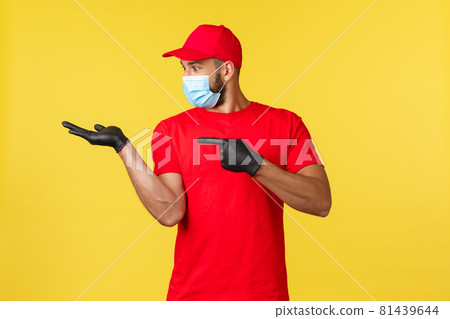 Express delivery during pandemic, covid-19, safe shipping, online shopping concept. Excited happy courier in red uniform and medical mask, gloves, pointing finger and looking at palm with product 81439644