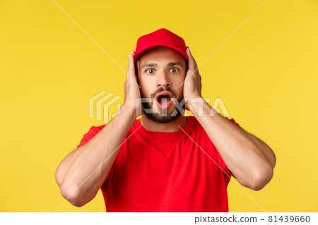 Close-up of shocked gasping male courier, employee in red uniform cap and t-shirt, cover ears and staring speechless at camera, heard stunning news, standing yellow background 81439660