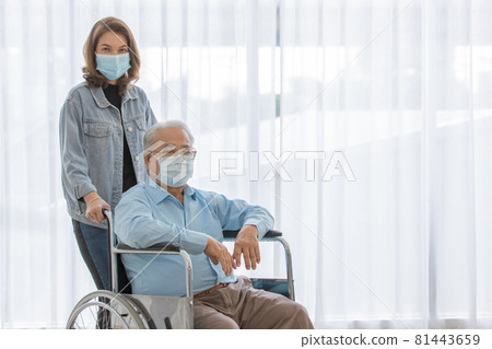 An old fat asian male patient with gray hair wearing light blue shirt and brown pants sitting on wheelchair when his daughter wearing jeans shirt stand holding the handles look at camera together An old fat asian male patient with gray hair wearing light blue shirt and brown pants sitting on wheelchair when his daughter wearing jeans shirt stand holding the handles look at camera together 81443659
