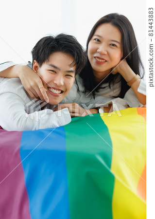 LGBTQ couple lovers, a handsome girl as a man or butch femme girl laying on a bed with the rainbow flag the symbol of LGBT, spending and sharing loving time in white bedroom with fun, warmth, and LGBTQ couple lovers, a handsome girl as a man or butch femme girl laying on a bed with the rainbow flag the symbol of LGBT, spending and sharing loving time in white bedroom with fun, warmth, and 81443883