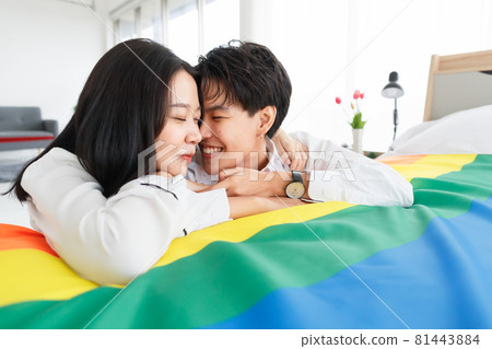 LGBTQ couple lovers, a handsome girl as a man or butch femme girl laying on a bed with the rainbow flag the symbol of LGBT, spending and sharing loving time in white bedroom with fun, warmth, and LGBTQ couple lovers, a handsome girl as a man or butch femme girl laying on a bed with the rainbow flag the symbol of LGBT, spending and sharing loving time in white bedroom with fun, warmth, and 81443884