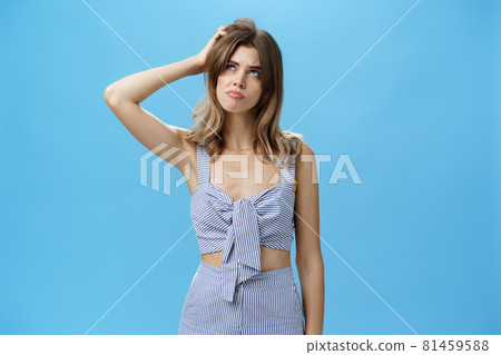 Silly confused charming adult female with chestnut hair in trendy outfit sulking looking questioned up with smirk and gloomy expression thinking scarthing head clueless over blue background 81459588