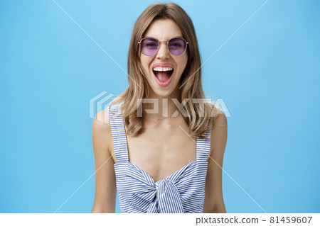 Expressive and careree woman being wild ad free yelling out loud at camera wearing awesome stylish sunglasses and striped top letting out emotions being open-minded and rebellious over blue wall Expressive and careree woman being wild ad free yelling out loud at camera wearing awesome stylish sunglasses and striped top letting out emotions being open-minded and rebellious over blue wall 81459607