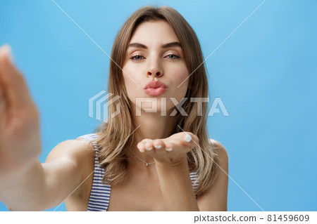 Close-up shot of self-assured glamorous and attractive stylish woman holding hand near folded lips to send kiss while grabbing camera and taking selfie standing sensual and romantic over blue wall Close-up shot of self-assured glamorous and attractive stylish woman holding hand near folded lips to send kiss while grabbing camera and taking selfie standing sensual and romantic over blue wall 81459609