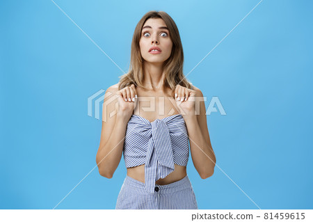 Portrait of stunned speechless and concerned young glamorous female pressing hands to chest popping eyes at camera standing in intense awkward and frightened pose being in stupor against blue wall Portrait of stunned speechless and concerned young glamorous female pressing hands to chest popping eyes at camera standing in intense awkward and frightened pose being in stupor against blue wall 81459615