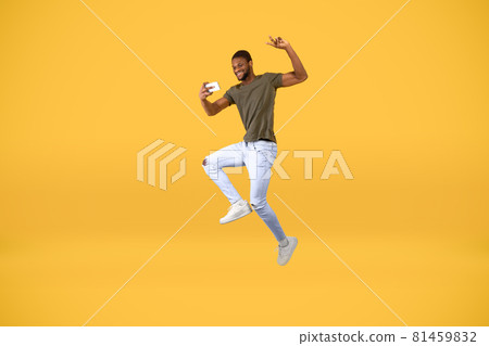 Crazy african american man making selfie on smartphone while jumping in the air over yellow studio background 81459832