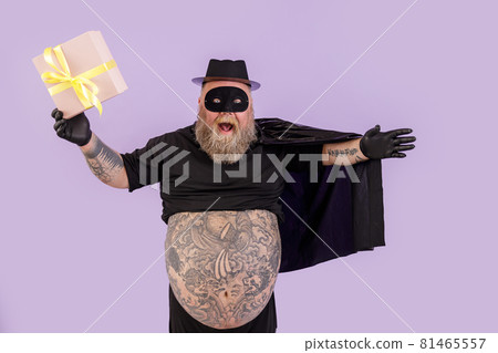 Joyful man with overweight in hero costume with cape holds gift on purple background Joyful man with overweight in hero costume with cape holds gift on purple background 81465557