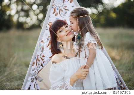 Cute girl with feathers in blond hair kisses her smiling cheerful mother. Boho mom and daughter Cute girl with feathers in blond hair kisses her smiling cheerful mother. Boho mom and daughter 81467301
