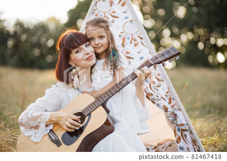 Hippie woman sits on the grass near teepee tent outdoors, plays guitar for her cute little daughte Hippie woman sits on the grass near teepee tent outdoors, plays guitar for her cute little daughte 81467418