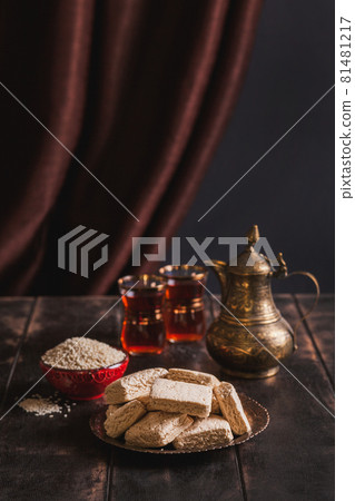 Pieces of tahini halva, sesame seeds in a bowl, tea in Turkish armudas, an vintage jug. Still life in oriental style on old brown wooden background Pieces of tahini halva, sesame seeds in a bowl, tea in Turkish armudas, an vintage jug. Still life in oriental style on old brown wooden background 81481217
