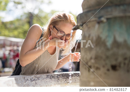 Thirsty young casual cucasian woman wearing medical face mask drinking water from public city fountain on a hot summer day. New social norms during covid epidemic Thirsty young casual cucasian woman wearing medical face mask drinking water from public city fountain on a hot summer day. New social norms during covid epidemic 81482817