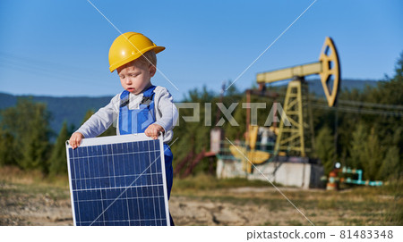 Child guy standing on blurred background of oil rocking pump and holding in his hands small solar panel. Future power engineer demonstrating ecological way of obtaining energy. Child guy standing on blurred background of oil rocking pump and holding in his hands small solar panel. Future power engineer demonstrating ecological way of obtaining energy. 81483348
