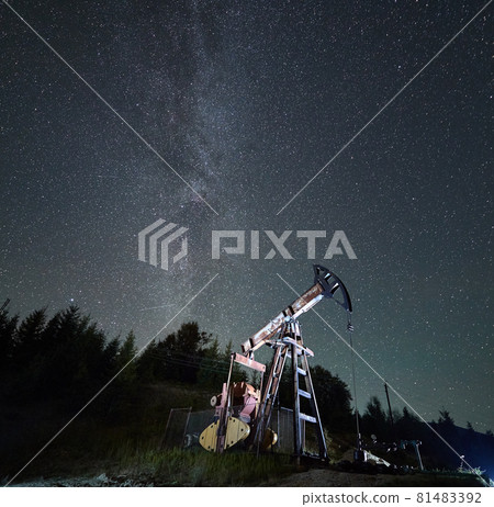 Special equipment for petroleum production. Oil derrick pump working at night. On the backdrop breathtaking scenery of starry sky, Milky Way galaxy and trees silhouettes. Special equipment for petroleum production. Oil derrick pump working at night. On the backdrop breathtaking scenery of starry sky, Milky Way galaxy and trees silhouettes. 81483392