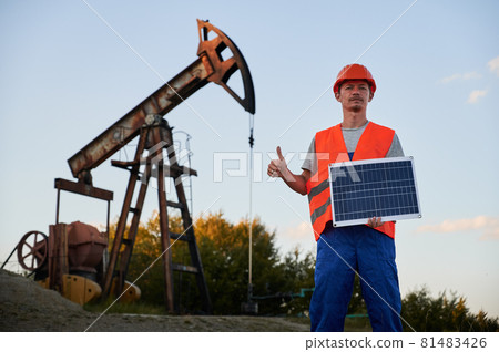 Portrait of serious engineer with solar panel in hand who showing class on the background of oil pump rig under blue sky. Environment friendly fotovoltaic energy conception. Portrait of serious engineer with solar panel in hand who showing class on the background of oil pump rig under blue sky. Environment friendly fotovoltaic energy conception. 81483426