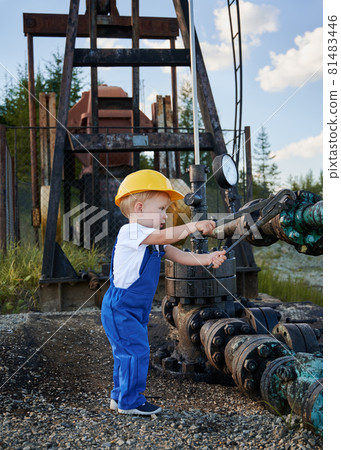 Side view of little boy in protective construction helmet who examining adjustable wrench by attaching it to nut on oil pipeline. On the background cropped view of pump jack. 81483446