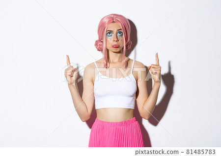 Portrait of miserable cute party girl in pink wig, sobbing and pouting upset, looking disappointed and pointing fingers up, standing over white background Portrait of miserable cute party girl in pink wig, sobbing and pouting upset, looking disappointed and pointing fingers up, standing over white background 81485783