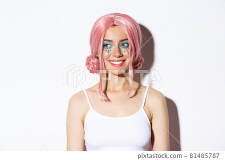 Close-up of lovely caucasian girl in pink wig, with bright makeup, looking left at logo and smiling, standing over white background 81485787