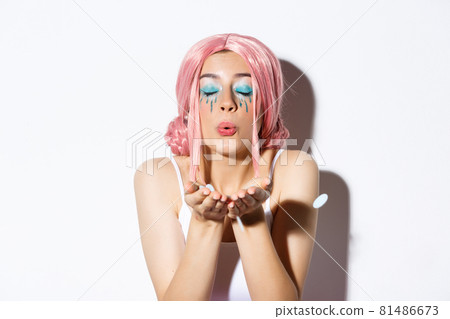 Close-up of lovely pretty party girl with pink wig and bright makeup, blowing confetti from palm, celebrating holiday, standing over white background 81486673