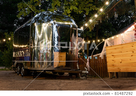 Scenic evening view of vintage futuristic silver metal camper van trailer parked near wooden fence at countryside farm or ranch against light garland forest tree. Retro style fodtruck caravan vehicle 81487292