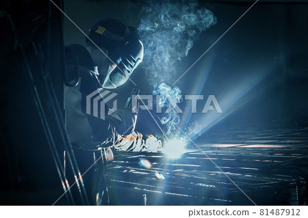 A masked working man is doing welding work on metal structures in a factory or industrial enterprise A masked working man is doing welding work on metal structures in a factory or industrial enterprise 81487912