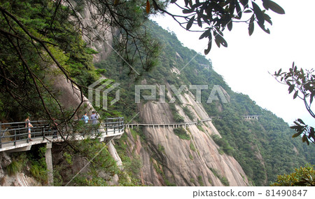 Sanqingshan Mountain in Jiangxi Province, China. People walking along a path clinging to the cliff high up on Mount Sanqing. Sanqingshan is a Taoist mountain famous for its peaks and forests. Sanqingshan Mountain in Jiangxi Province, China. People walking along a path clinging to the cliff high up on Mount Sanqing. Sanqingshan is a Taoist mountain famous for its peaks and forests. 81490847