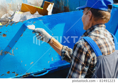 An old man in a work jumpsuit paints a garbage can or container on the street with a paint brush. An old man in a work jumpsuit paints a garbage can or container on the street with a paint brush. 81491223