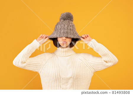 Pretty young woman pulling down woollen hat, showing kissing lips, fooling and posing to camera over yellow background 81493808