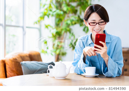 戴眼鏡的中年婦女在客廳喝茶時使用智能手機 81494390