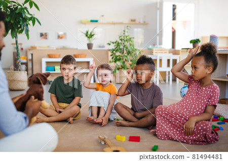 Group of small nursery school children with man teacher sitting on floor indoors in classroom, raising hands. Group of small nursery school children with man teacher sitting on floor indoors in classroom, raising hands. 81494481