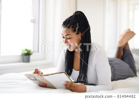 Happy teenage girl student lying indoors on bed at home, reading book and making notes. 81494482