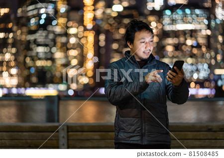 Asian man wearing overcoat suit using smart mobile phone with smile action over the photo blurred bokeh of New york Cityscape beside the east river, USA downtown, career working everywhere concept 81508485