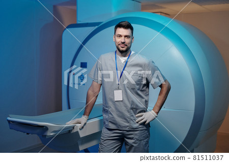 Confident clinician in grey uniform standing by table of mri scan equipment 81511037