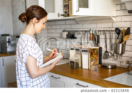 Smiling young beautiful business female taking notes in paper diary at modern Nordic style kitchen 81513848