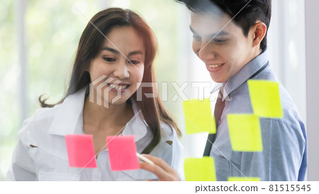 Beautiful black long hair Asian businesswoman and gook looking cute Asian businessman join together in discussion and brainstorming, they write the idea on colorful memo paper on glass board 81515545