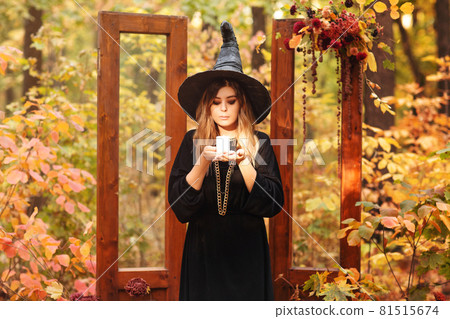 Young woman in Witch costum and hat with candle in hands stands against autumn forest background 81515674