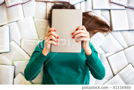 Brunette woman in green sweater lies on large number of open books while holding digital tablet Brunette woman in green sweater lies on large number of open books while holding digital tablet 81516980