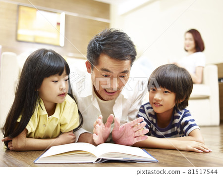 asian father and two children lying on front on floor reading book mother watching in background asian father and two children lying on front on floor reading book mother watching in background 81517544
