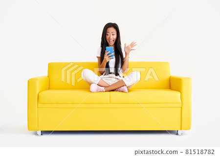 Emotional surprised asian female receiving good message on cell phone sitting on yellow couch Emotional surprised asian female receiving good message on cell phone sitting on yellow couch 81518782