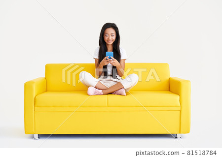 Smiling asian woman types text message, browses internet on cell phone sitting on yellow couch Smiling asian woman types text message, browses internet on cell phone sitting on yellow couch 81518784