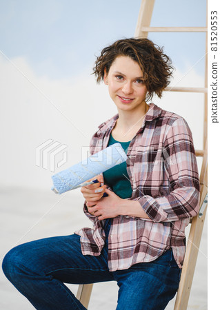 A young beautiful woman painter designer paints a white wall with blue paint with roller. Plaid shirt, short hair and jeans. 81520553