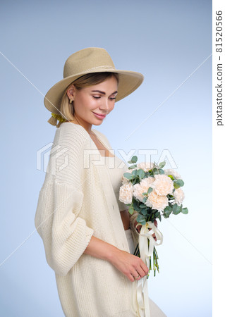 A young beautiful blonde caucasian woman with a short haircut in a beige suit and hat with bouquet of white flowers on blue background. Beauty portrait. The girl smiles, clean skin and white teeth. A young beautiful blonde caucasian woman with a short haircut in a beige suit and hat with bouquet of white flowers on blue background. Beauty portrait. The girl smiles, clean skin and white teeth. 81520566