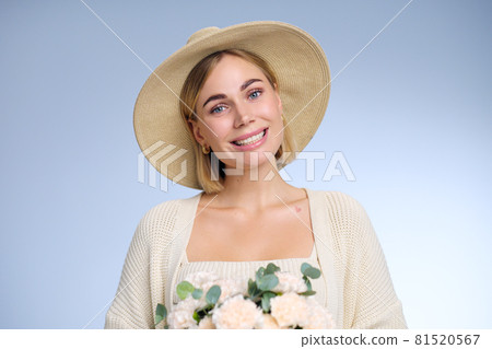 A young beautiful blonde caucasian woman with a short haircut in a beige suit and hat with bouquet of white flowers on blue background. Beauty portrait. The girl smiles, clean skin and white teeth. A young beautiful blonde caucasian woman with a short haircut in a beige suit and hat with bouquet of white flowers on blue background. Beauty portrait. The girl smiles, clean skin and white teeth. 81520567