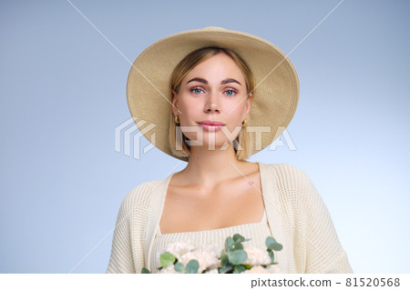 A young beautiful blonde caucasian woman with a short haircut in a beige suit and hat with bouquet of white flowers on blue background. Beauty portrait. The girl smiles, clean skin and white teeth. A young beautiful blonde caucasian woman with a short haircut in a beige suit and hat with bouquet of white flowers on blue background. Beauty portrait. The girl smiles, clean skin and white teeth. 81520568