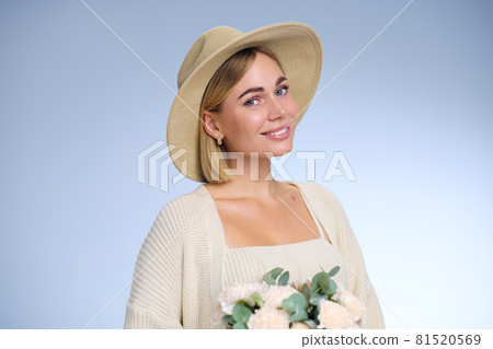 A young beautiful blonde caucasian woman with a short haircut in a beige suit and hat with bouquet of white flowers on blue background. Beauty portrait. The girl smiles, clean skin and white teeth. A young beautiful blonde caucasian woman with a short haircut in a beige suit and hat with bouquet of white flowers on blue background. Beauty portrait. The girl smiles, clean skin and white teeth. 81520569