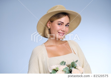 A young beautiful blonde caucasian woman with a short haircut in a beige suit and hat with bouquet of white flowers on blue background. Beauty portrait. The girl smiles, clean skin and white teeth. A young beautiful blonde caucasian woman with a short haircut in a beige suit and hat with bouquet of white flowers on blue background. Beauty portrait. The girl smiles, clean skin and white teeth. 81520575