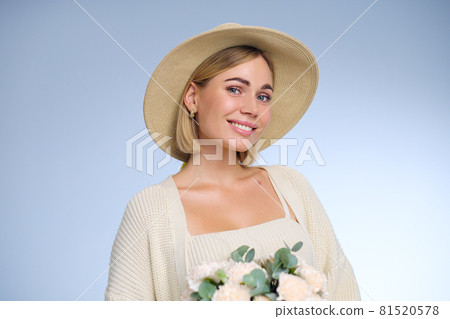 A young beautiful blonde caucasian woman with a short haircut in a beige suit and hat with bouquet of white flowers on blue background. Beauty portrait. The girl smiles, clean skin and white teeth. A young beautiful blonde caucasian woman with a short haircut in a beige suit and hat with bouquet of white flowers on blue background. Beauty portrait. The girl smiles, clean skin and white teeth. 81520578